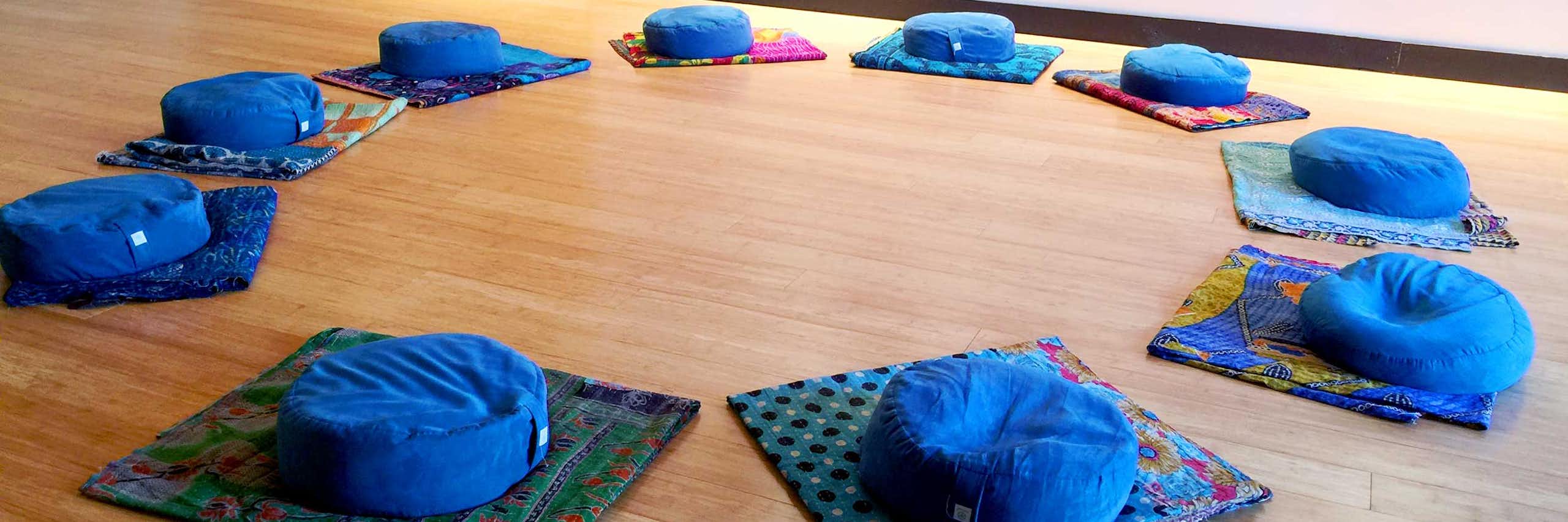 Several blue cushions are arranged in a circle on several mats laid out on a wooden floor.