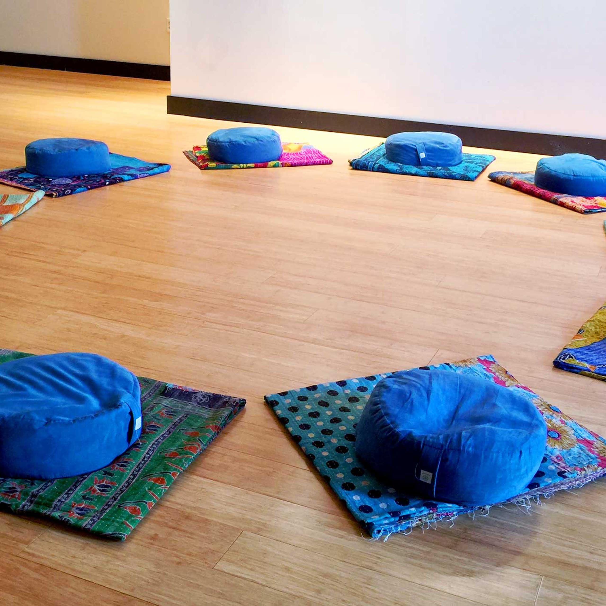 Several blue cushions are arranged in a circle on several mats laid out on a wooden floor.