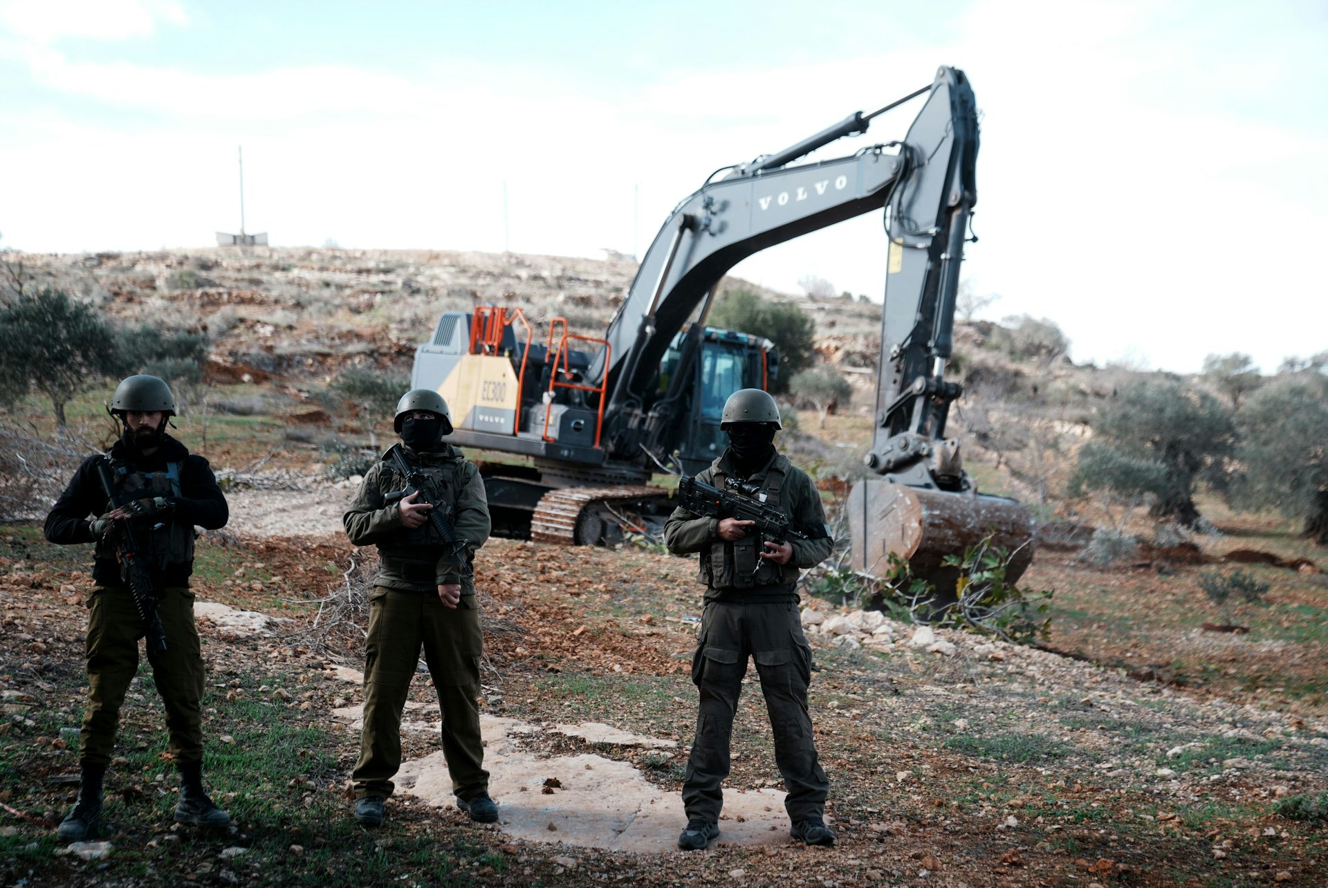 Three soldiers stand in front of a mechanical digger