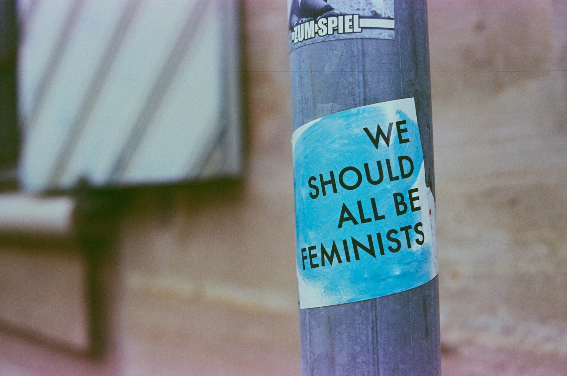 A sticker that reads "We should all be feminists" is posted on a street pole.