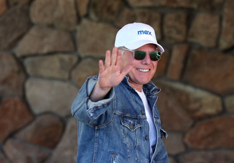 Whether or not Netflix or Paramount buys Warner Bros., leisure oligopolies are again – larger and extra anticompetitive than ever 1 Older man smiles and waves while wearing sunglasses and a white baseball cap reading 'Max.'