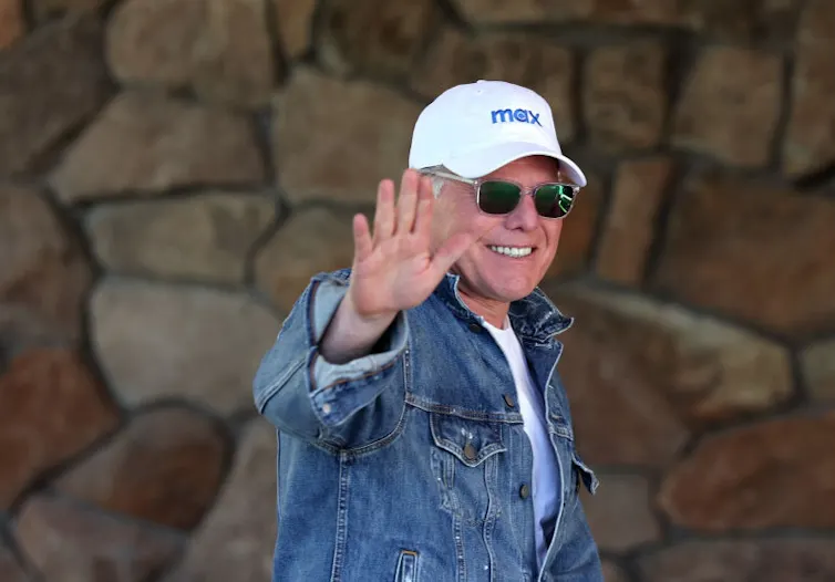 Older man smiles and waves while wearing sunglasses and a white baseball cap reading 'Max.'