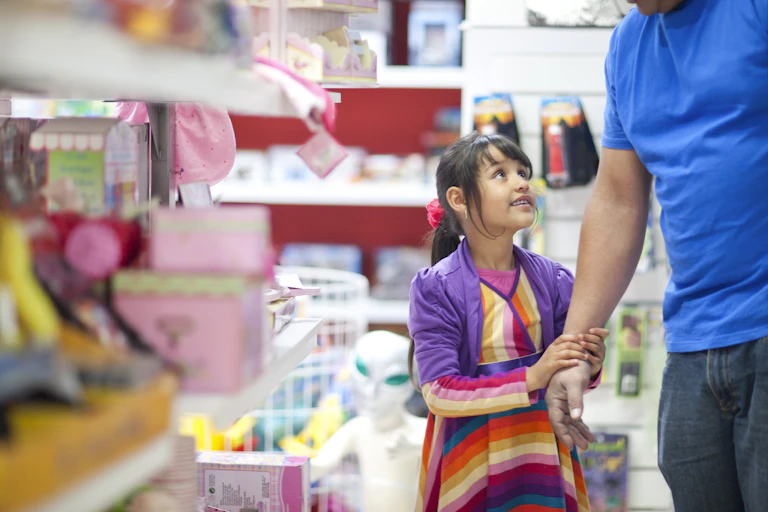 Uma menina parada em um corredor de brinquedos, vestindo roupas coloridas, olha para um homem adulto de camisa azul segurando sua mão.