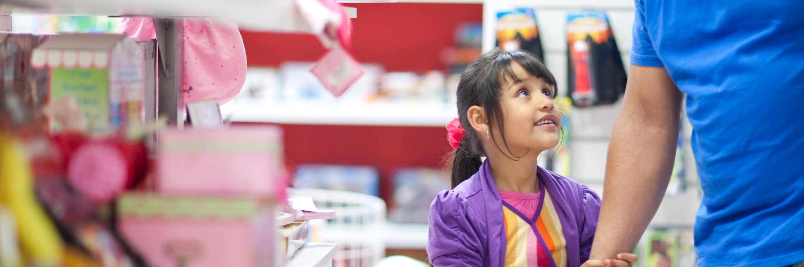 Uma menina parada em um corredor de brinquedos, vestindo roupas coloridas, olha para um homem adulto de camisa azul segurando sua mão.