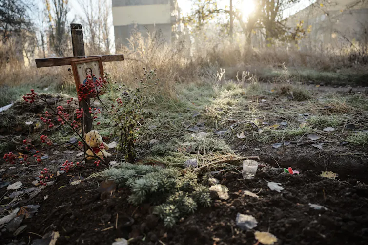Sunlight streams over an overgrown yard with a simple wooden cross and a small teddy bear.
