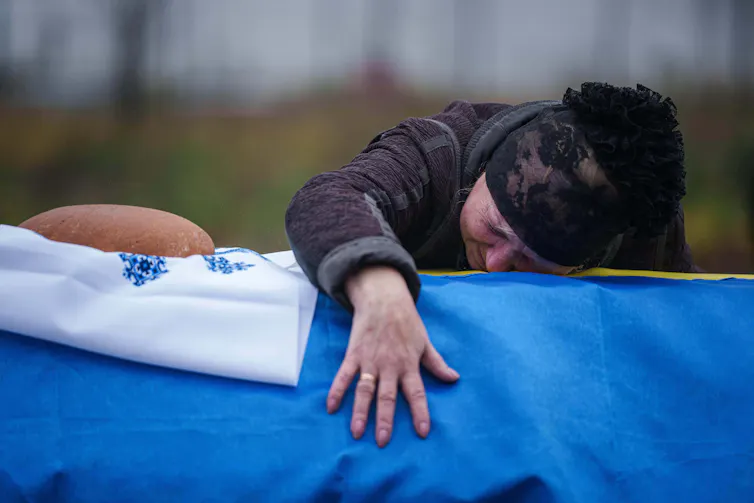 A woman in black clothes and a black hat leans over a coffin draped in sky blue fabric.