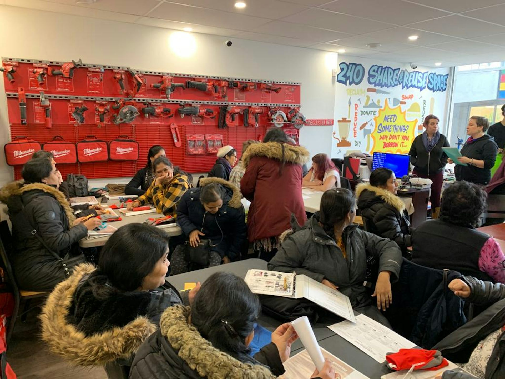 People in a room sit at desks listening to a standing speaker