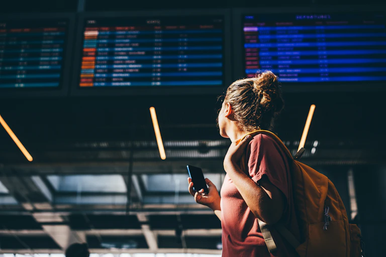 Young woman, carrying bag and phone, at airport looking up at departure board.