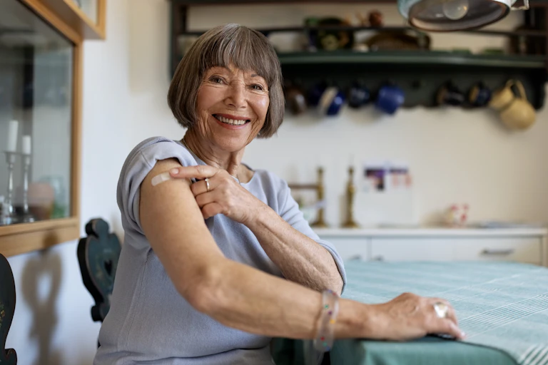 Older woman sitting down in kitchen pointing to bandaid on upper arm and smiling