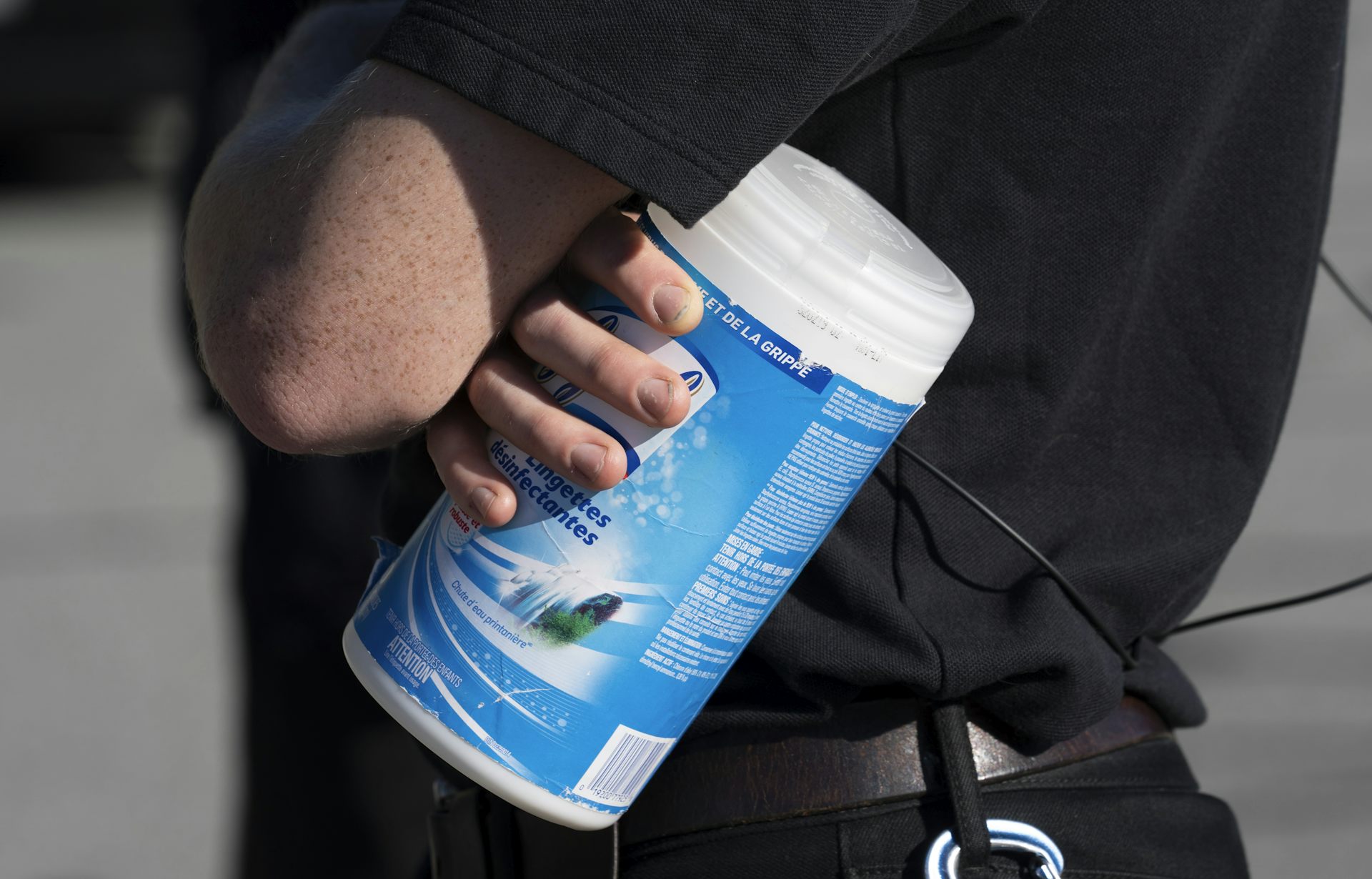 Foto do torso de uma pessoa com roupas escuras, segurando uma lata de lenços umedecidos desinfetantes