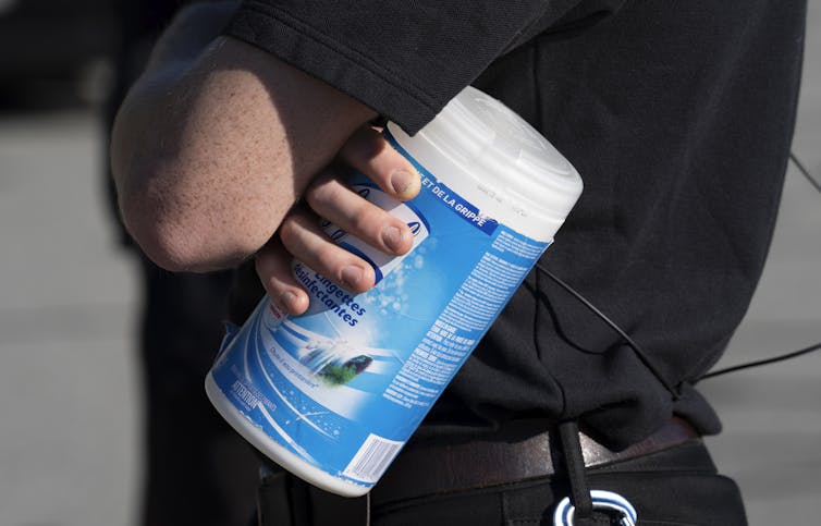 Foto do torso de uma pessoa com roupas escuras, segurando uma lata de lenços umedecidos desinfetantes