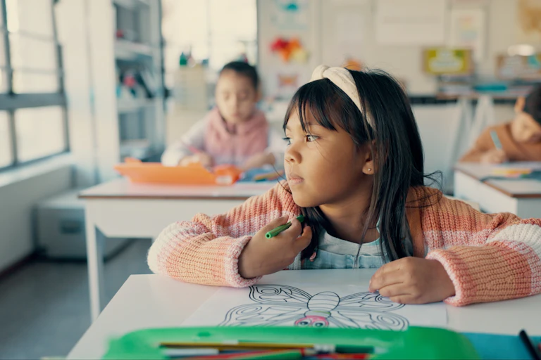 A girl sitting at her desk in school with a colouring page. She is staring out the window instead of colouring.