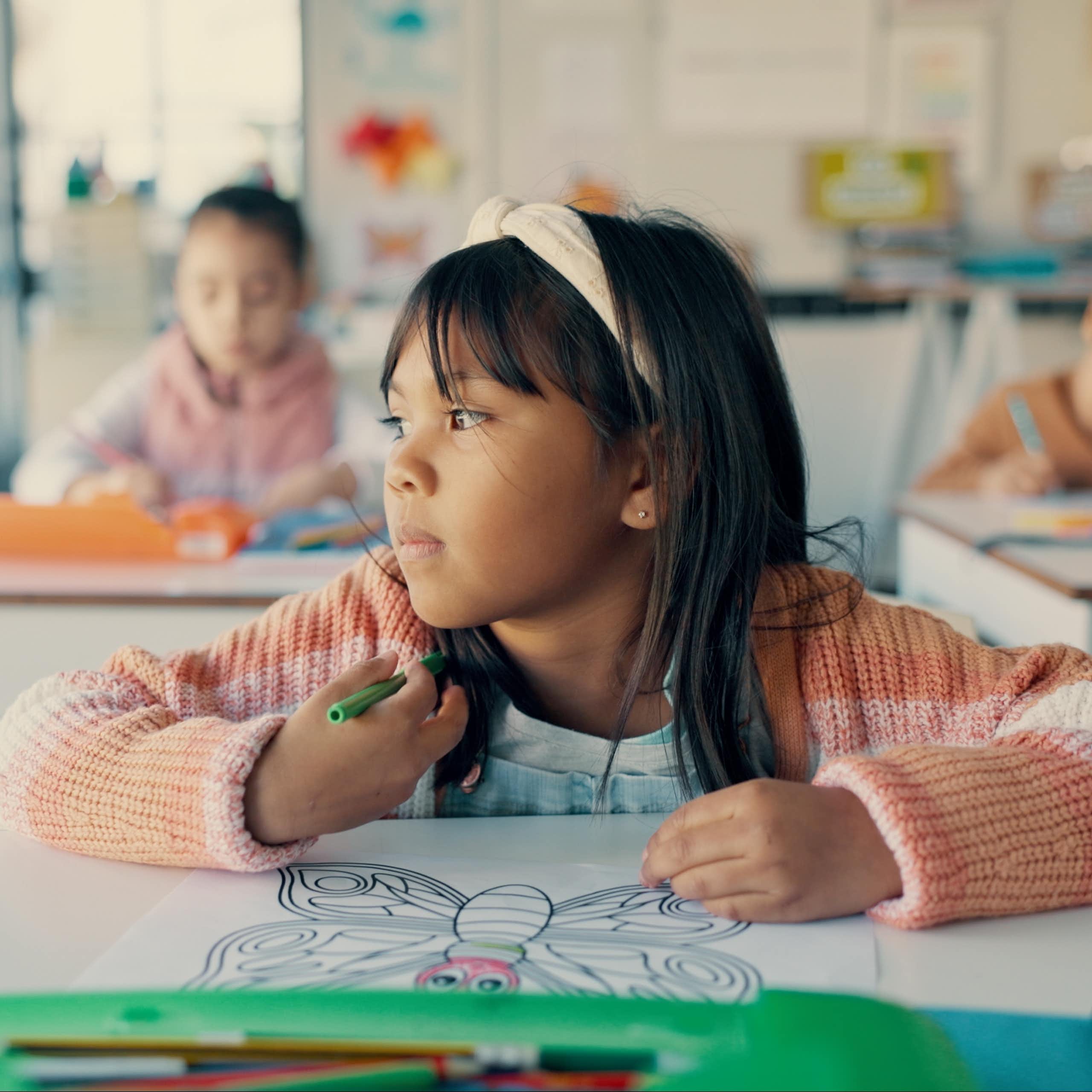 A girl sitting at her desk in school with a colouring page. She is staring out the window instead of colouring.