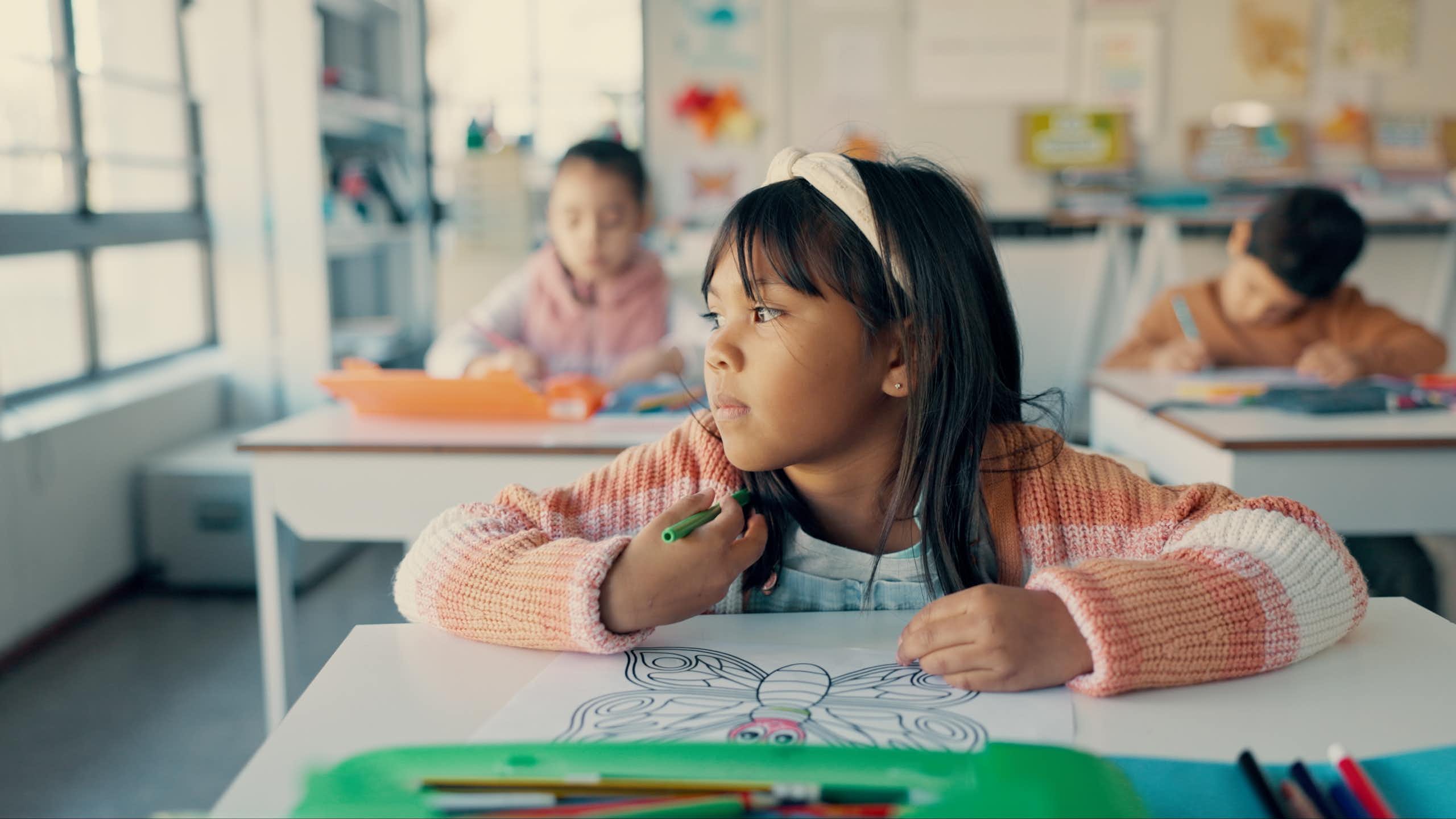 A girl sitting at her desk in school with a colouring page. She is staring out the window instead of colouring.
