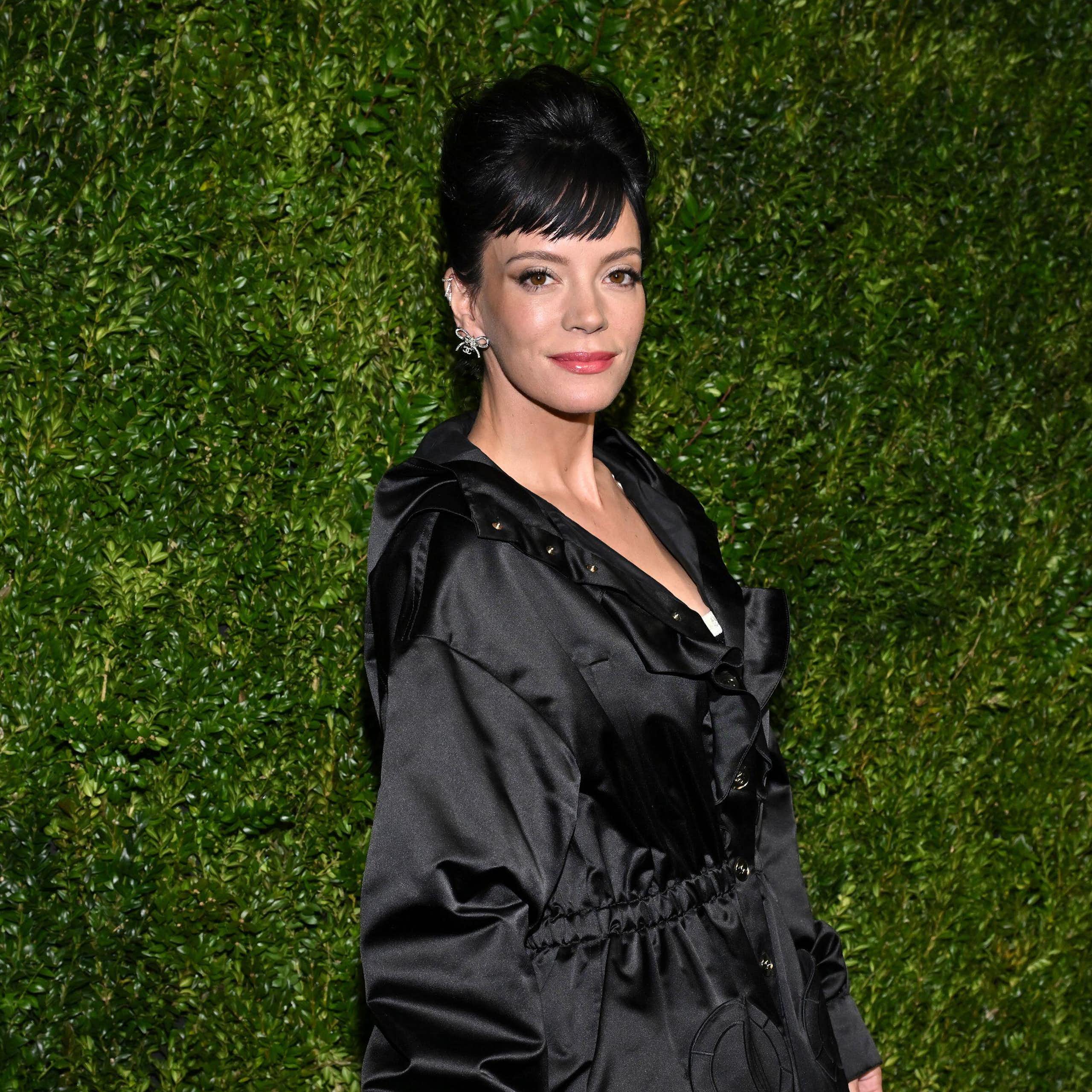 Lily Allen dressed in black for the red carpet against a background of green foliage.