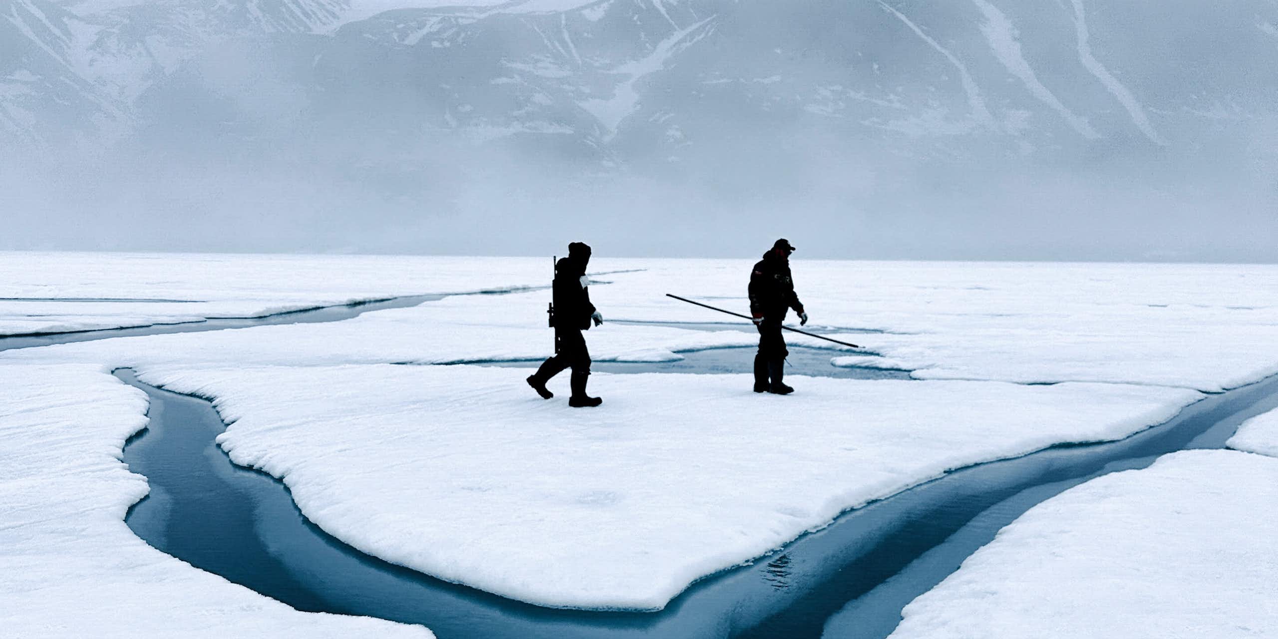 Two people walk across snow and ice with areas of water evident and mountains in the background.