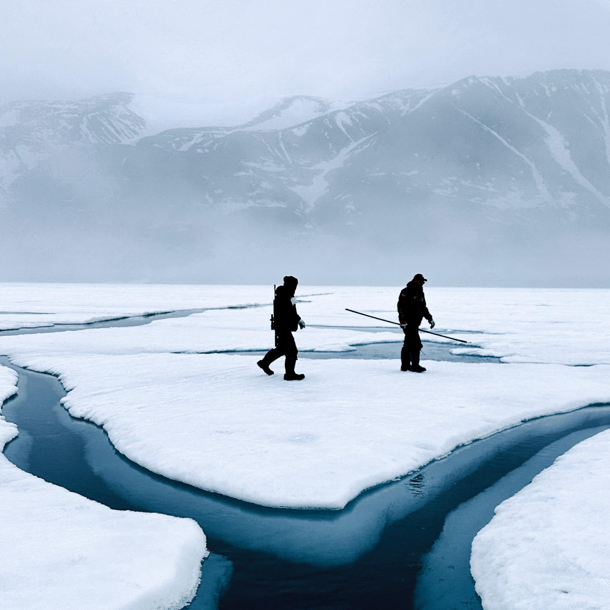 Two people walk across snow and ice with areas of water evident and mountains in the background.