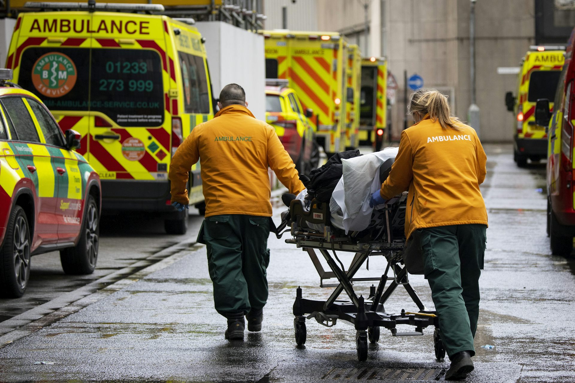 A patient being wheeled into hospital on a gurney. A queue of ambulances sits in the foreground.