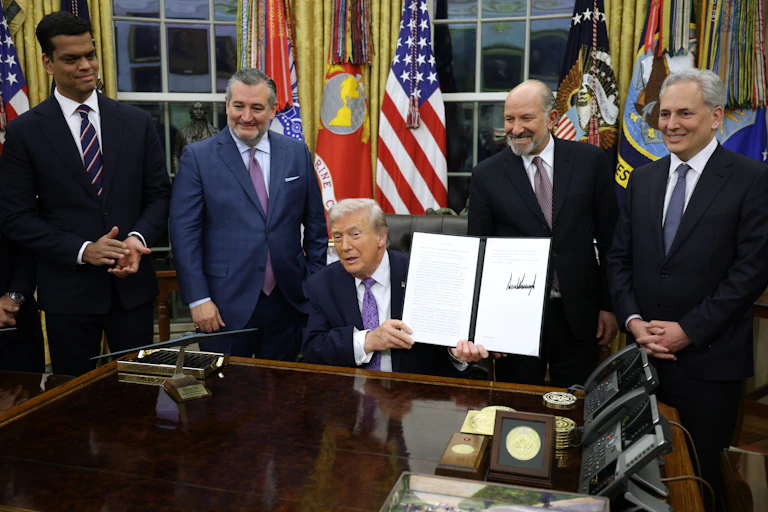 A man in a business suit seated at a desk holds up a document as other men in business suits stand behind him.