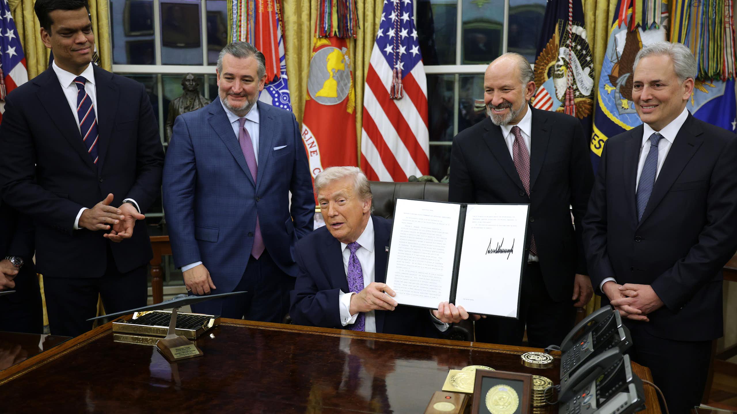 A man in a business suit seated at a desk holds up a document as other men in business suits stand behind him.