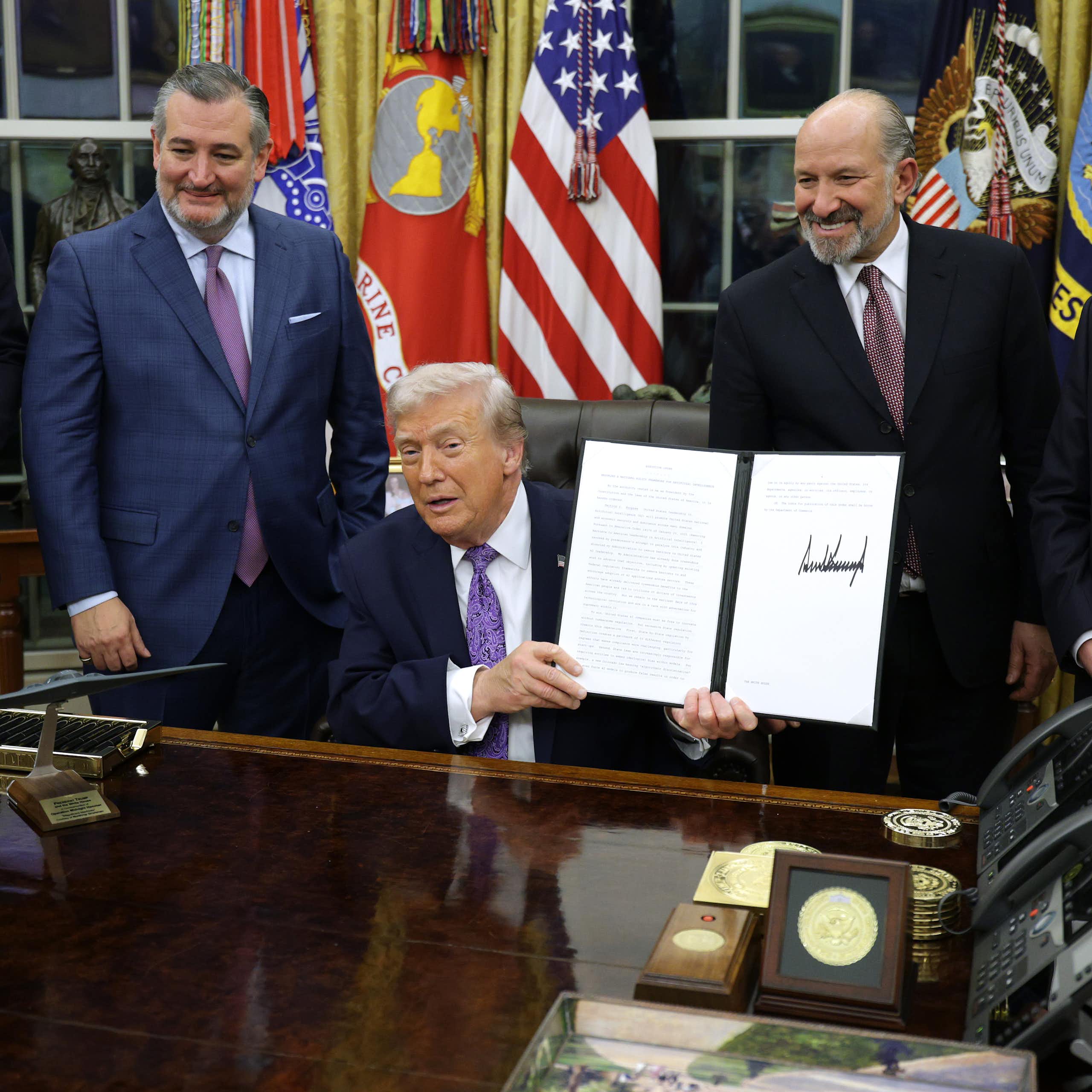 A man in a business suit seated at a desk holds up a document as other men in business suits stand behind him.