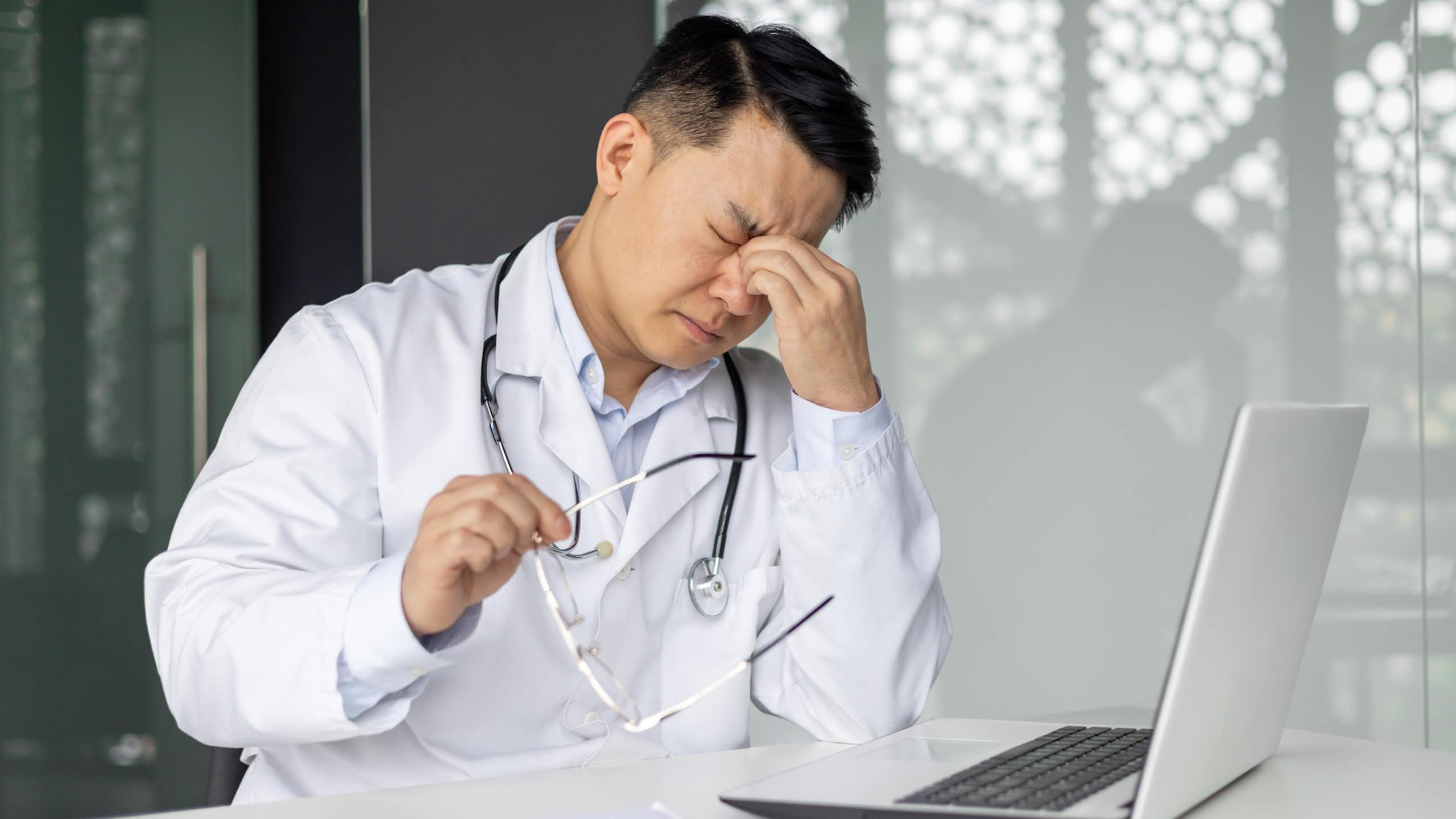 Doctor looking stressed while sitting at desk with laptop and holding glasses
