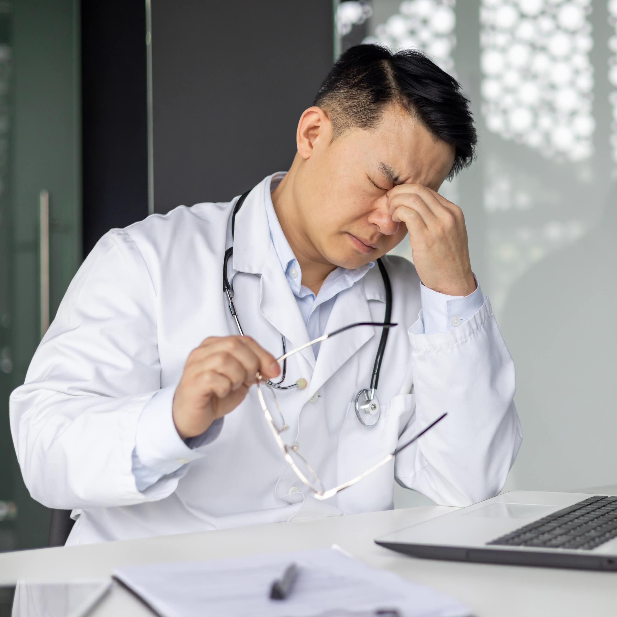 Doctor looking stressed while sitting at desk with laptop and holding glasses