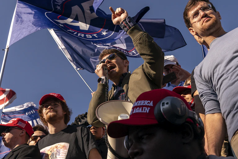 Young man wearing sunglasses shouting into a microphone surrounded by other young men.