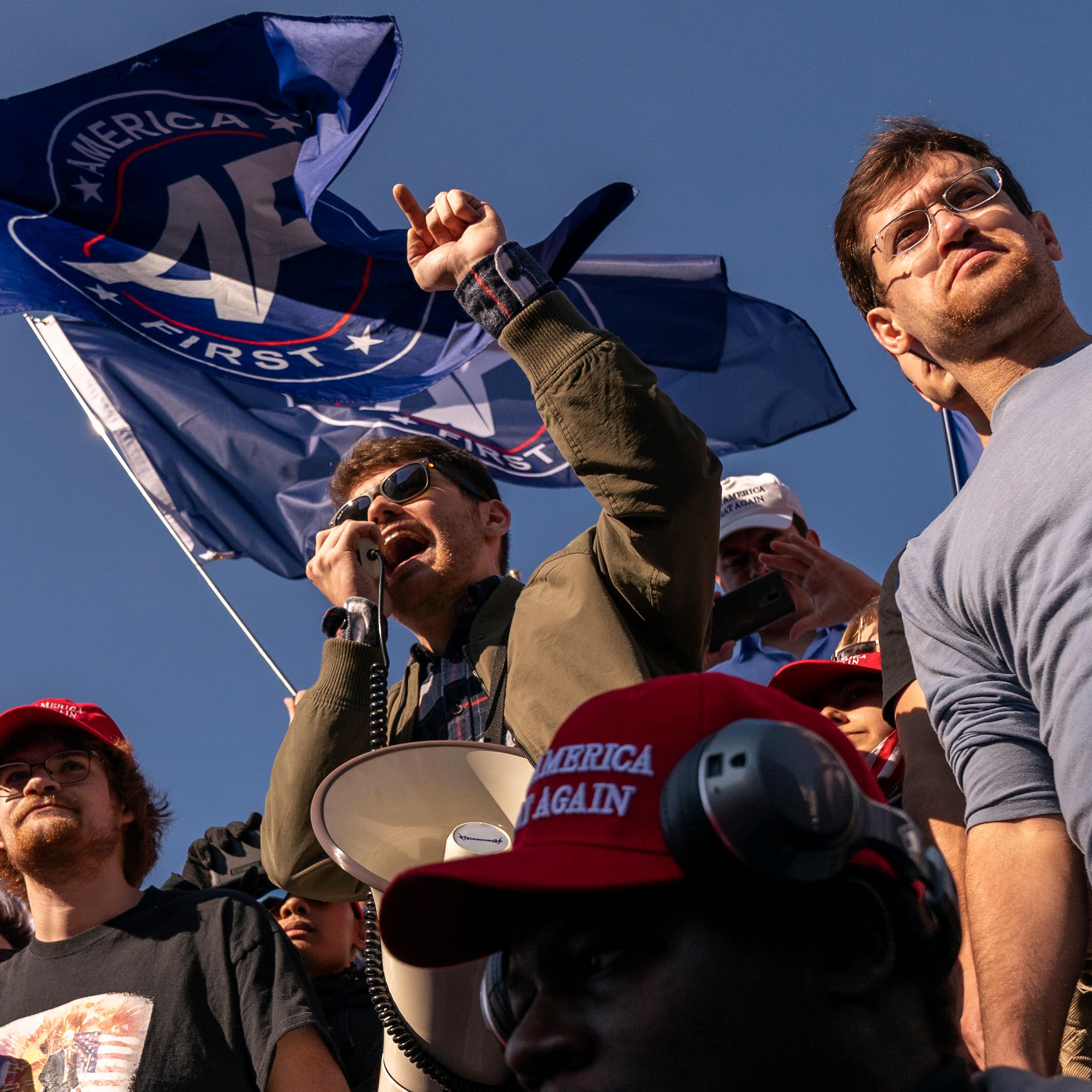 Young man wearing sunglasses shouting into a microphone surrounded by other young men.