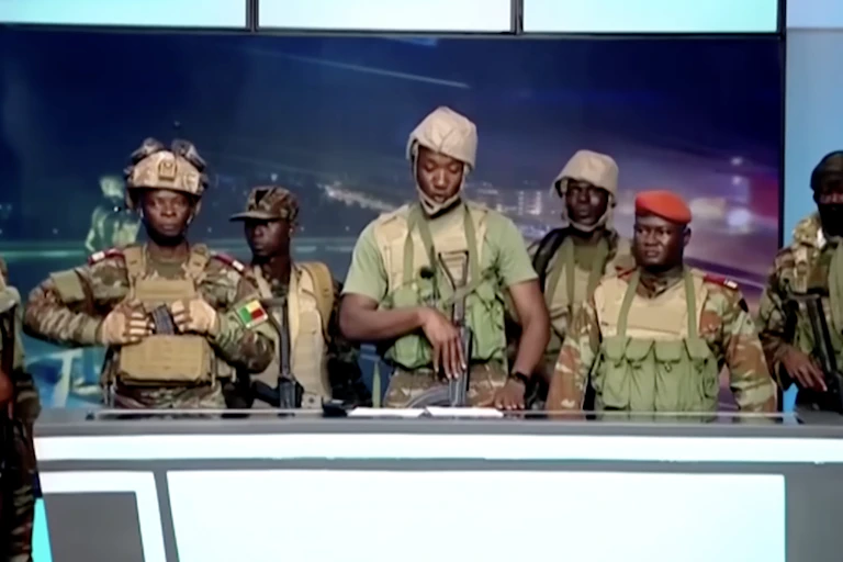 A group of men in army fatigues and holding guns stand behind a desk.
