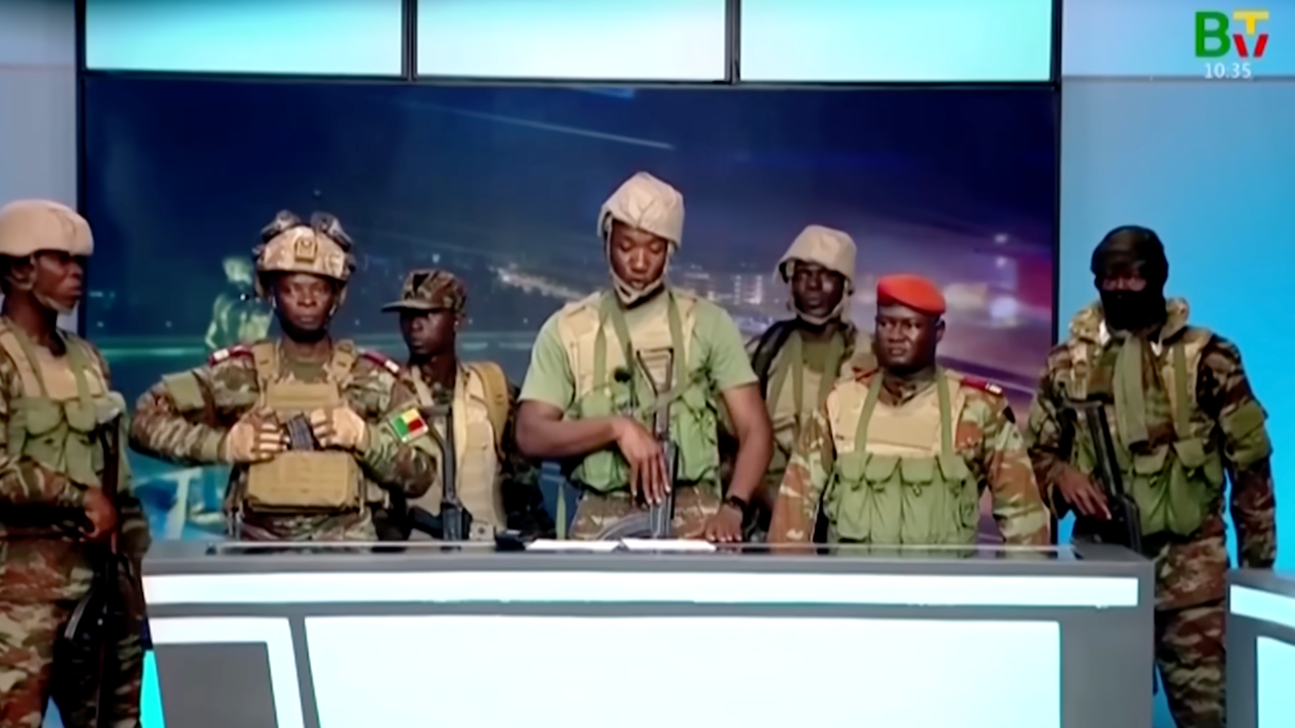 A group of men in army fatigues and holding guns stand behind a desk.