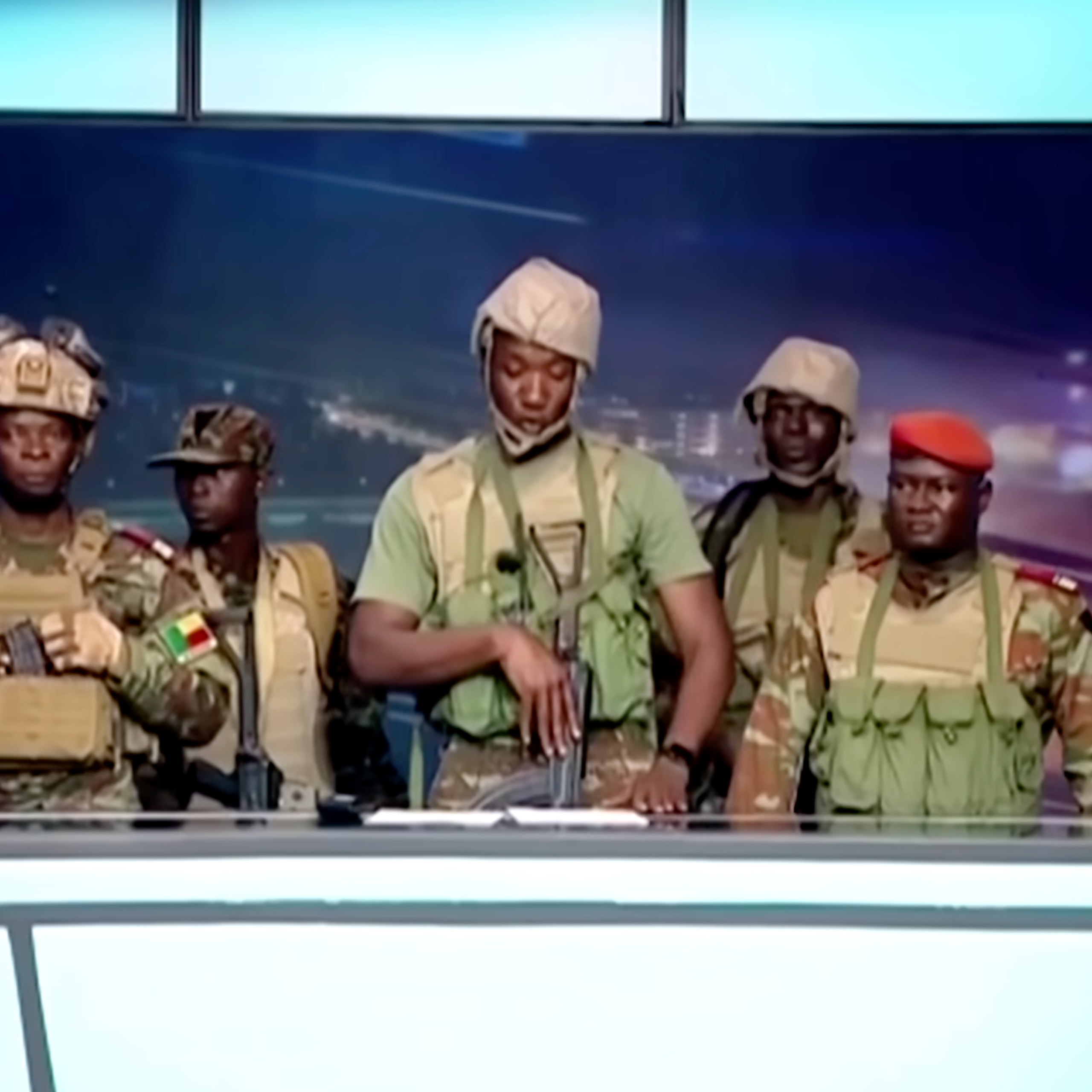 A group of men in army fatigues and holding guns stand behind a desk.