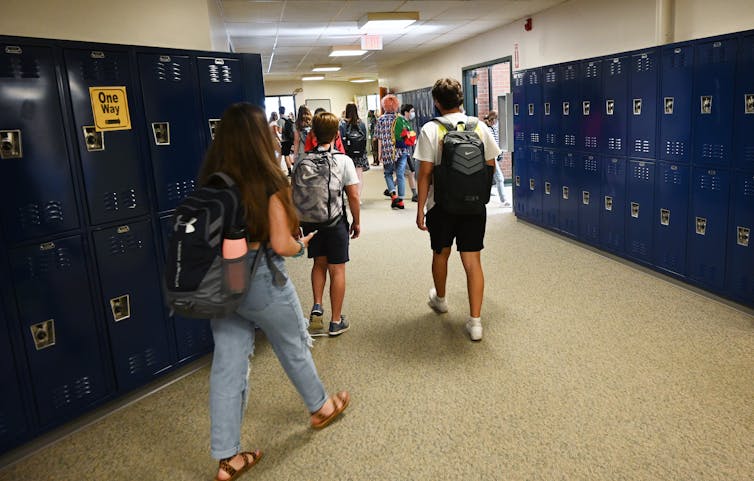 De espaldas se ve a un grupo de jóvenes caminando frente a los casilleros y cargando mochilas.