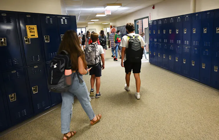 A group of young people are seen from behind walking in front of lockers and carrying backpacks.