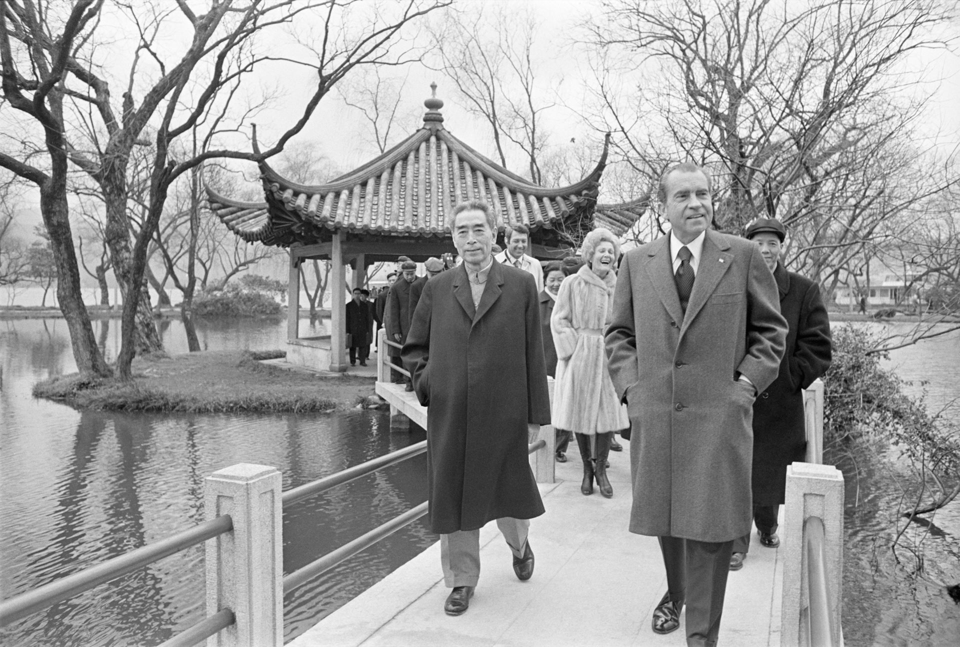 Two men in overcoats walk on a bridge in front of a pagoda.