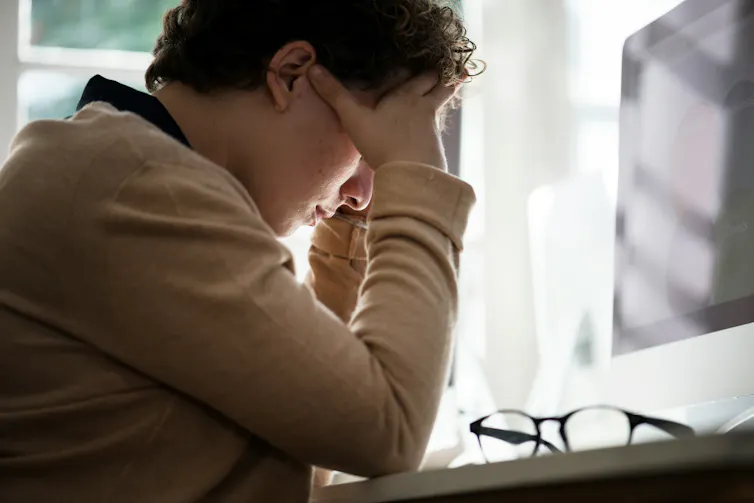A person sits at a computer desk with their head in their hands