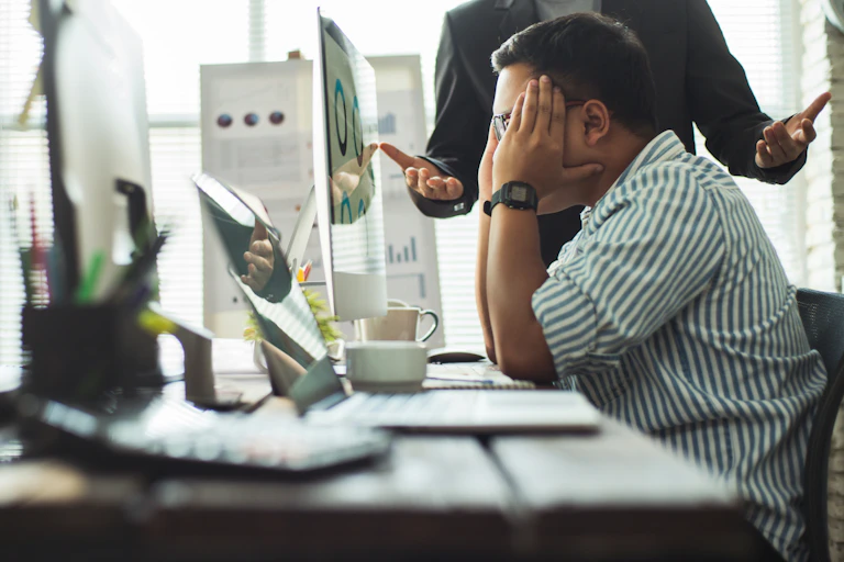 A person sits in front of a computer rubbing their temples while another person, seen from the shoulders down, gestures forcefully while speaking to them