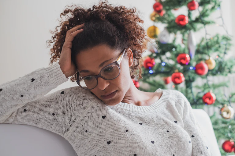 Woman sitting on sofa with Christmas tree behind her, looking dejected and holding her head with her hand