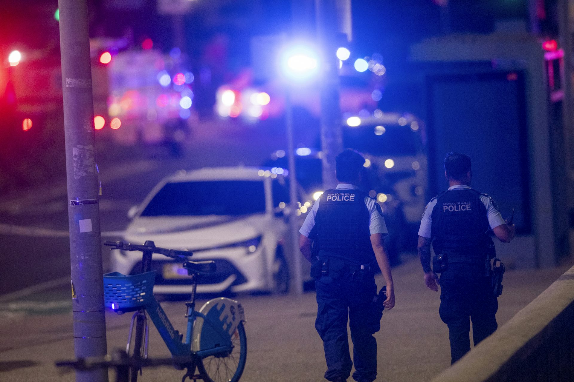 The backs of two police officers walking down a street lit up by police car lights