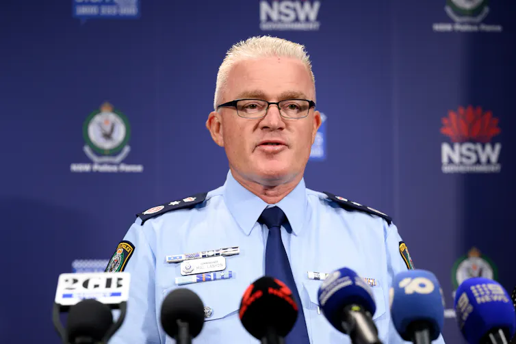 A man in a police shirt and tie speaks at a podium behind a row of microphones