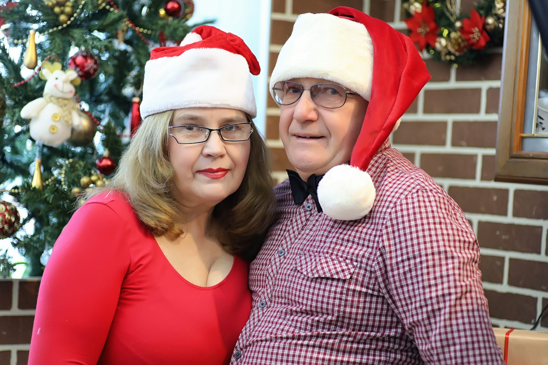 Middle-aged couple stand in front of Christmas tree wearing Santa hats and looking unimpressed