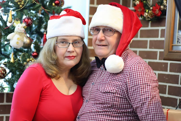 Middle-aged couple stand in front of Christmas tree wearing Santa hats and looking unimpressed