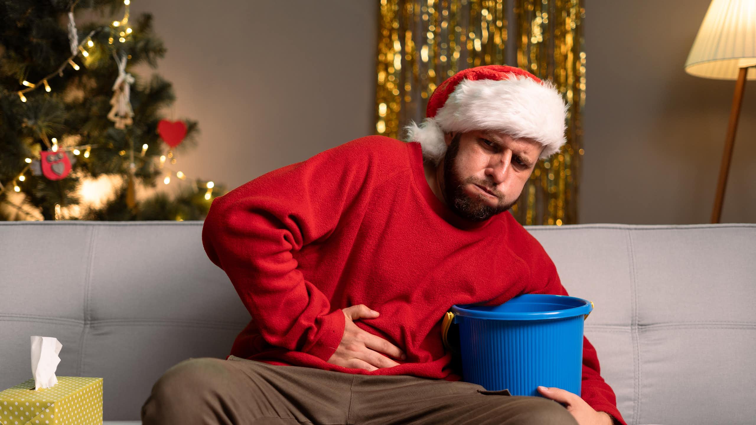 Man sitting on sofa in Christmas jumper and Santa hat with Christmas tree in background holds sick bucket in one hand and his stomach with the other