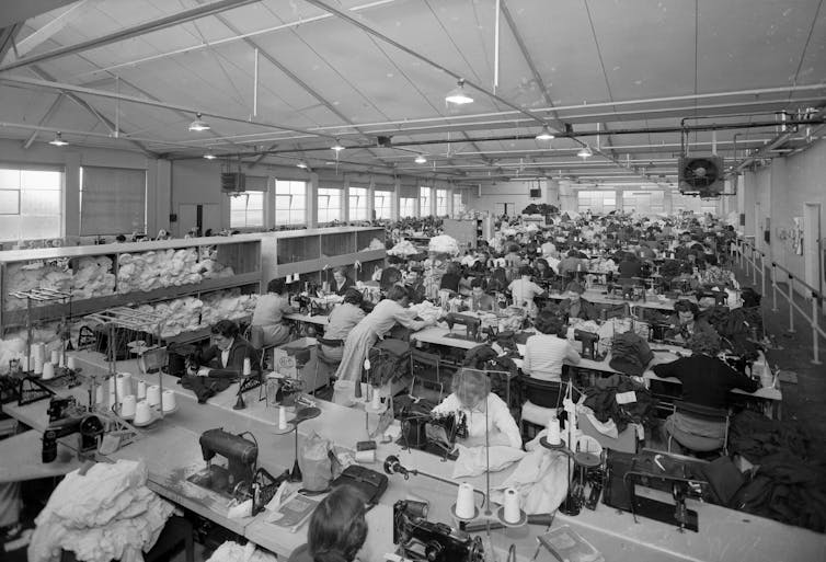 Uma imagem em preto e branco de um grande número de mulheres costurando roupas em um edifício industrial.