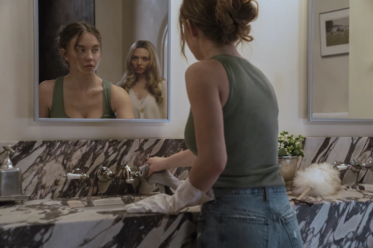 Two women reflected in a bathroom mirror, one cleans, one watches ominously