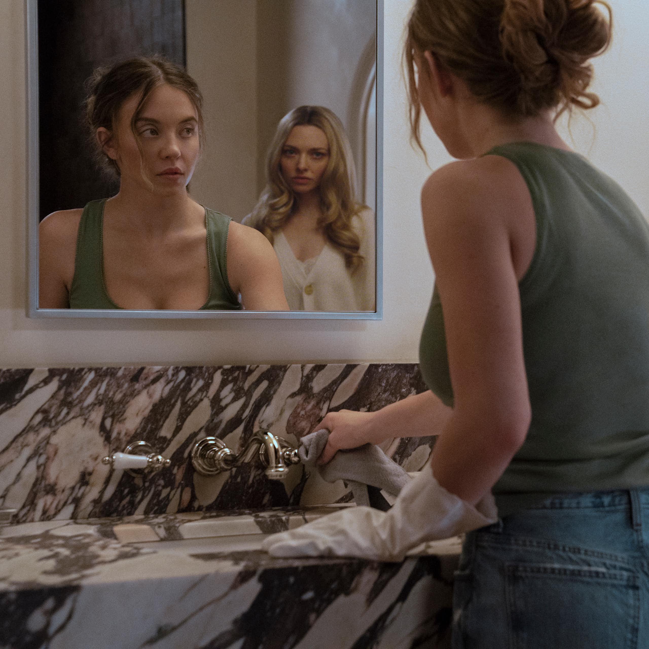 Two women reflected in a bathroom mirror, one cleans, one watches ominously