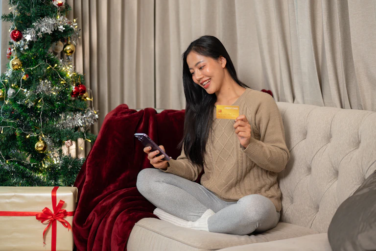 A woman is doing some online Christmas shopping on her phone while sitting on a sofa in the living room.