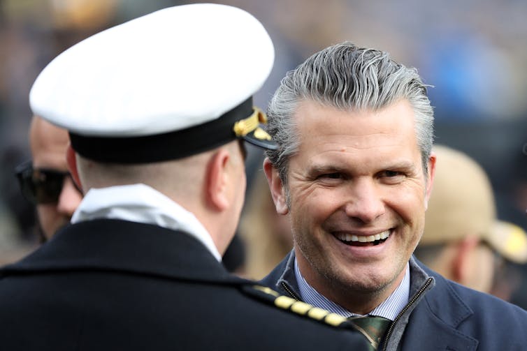 A man speaks to another man wearing a white cap.