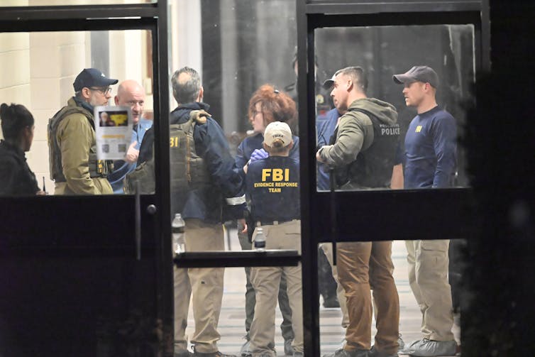 A group of officials wearing green and blue FBI and law enforcement shirts and vests stand inside a room, seen through glass doors with dark paneling.