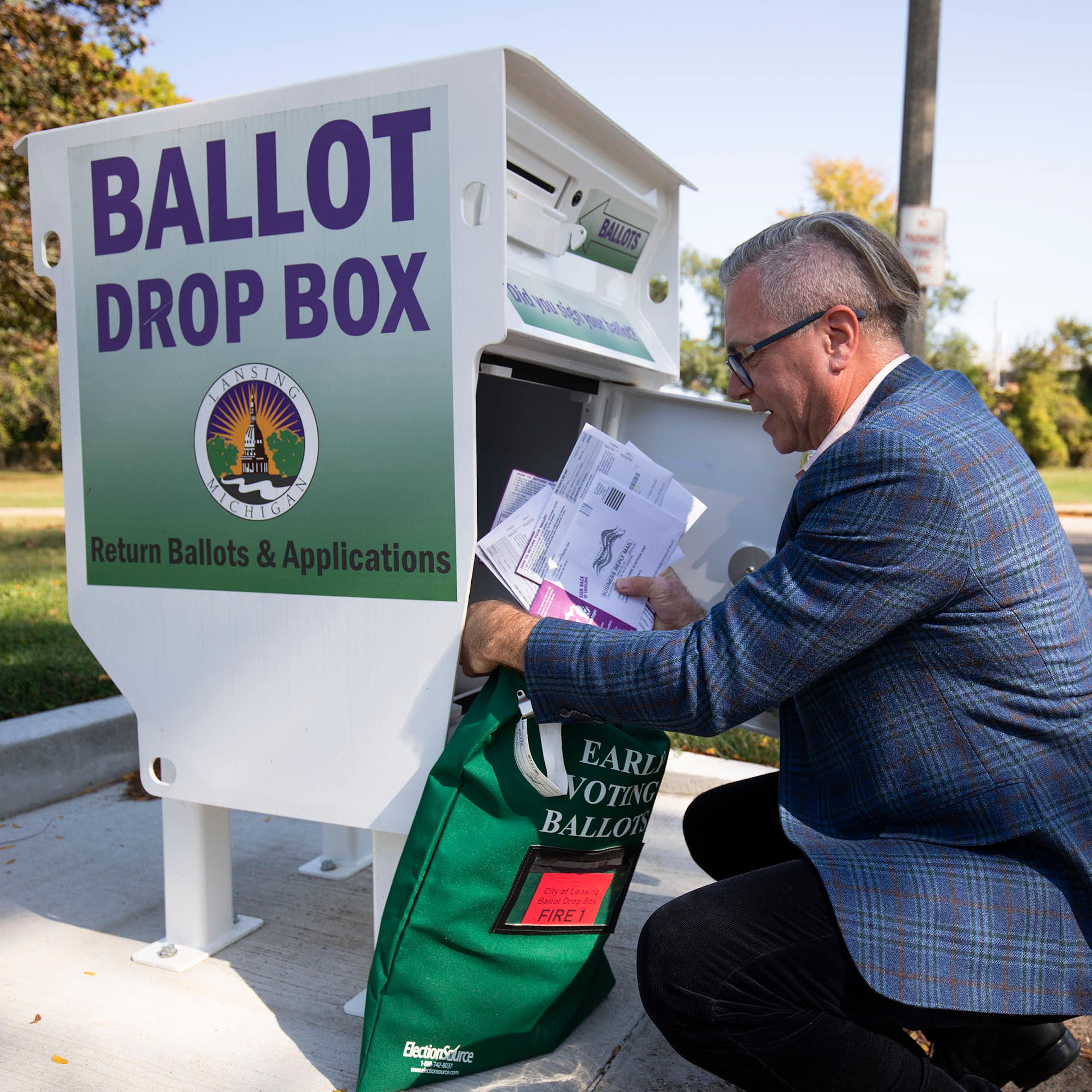 A man wearing glasses and a blue sports jacket kneels to remove ballots from a green box reading "BALLOT DROP BOX"